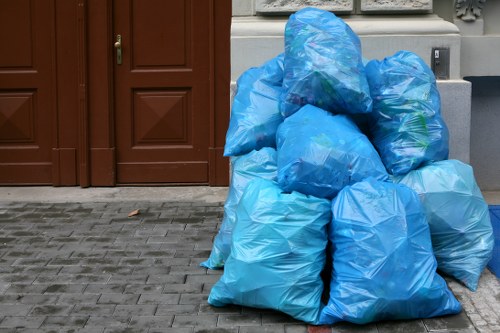 A group of securely tied, large blue plastic rubbish bags filled with waste are stacked and placed on a paved driveway in front of a building with brown wooden doors. The bags vary in size and are made of semi-transparent plastic, allowing some contents to be faintly visible through the material. They are positioned close together, occupying the lower right section of the image, with some leaning against each other for stability. The paving is composed of grey bricks arranged in a regular pattern, and part of a light-colored wall with decorative detailing is visible behind the bags. This scene illustrates typical rubbish collection or removal activities, possibly managed by local waste removal services such as Business Waste Removal Victoria, servicing the Victoria area or nearby postcode regions. The environment appears clean and orderly, suitable for commercial or residential waste disposal. The lighting is natural, with even illumination across the scene, emphasizing the colours and textures of the bags and surroundings.