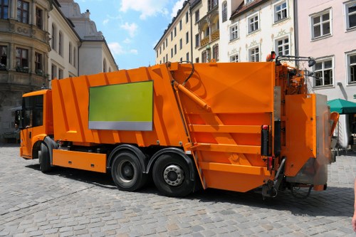 A large, bright orange municipal waste collection truck parked on a cobbled street in an urban area with historic European-style buildings in the background. The truck has a reflective green and white panel on its side, with visible hydraulic components at the rear used for waste compaction. The vehicle's textured metal surface is painted in a vivid orange finish, typical for city waste management vehicles, and it is positioned to the right of the image, occupying most of the scene. The setting appears to be a public square or pedestrianized street, with the cobblestone paving clearly visible beneath the truck. The sky overhead is partly cloudy, providing natural light that highlights the truck's reflective finish and detailed features. This image depicts a typical waste management activity likely associated with rubbish collection services offered by businesses such as Business Waste Removal Victoria, serving areas around the Victoria postcode in central London, where municipal waste collection and rubbish removal are part of local infrastructure.