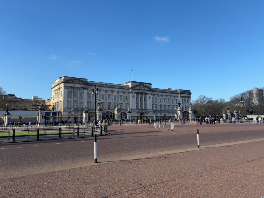 A wide, paved avenue lined with tall, leafy trees on both sides, featuring several flagpoles evenly spaced along the street. Each flagpole displays the Union Jack flag, fluttering gently in the breeze