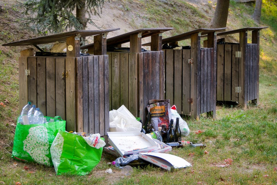 Four wooden outdoor rubbish bins with flat, sloped roofs are positioned in a row on a grassy area, partially surrounded by trees and foliage. The bins are made of vertical wooden planks with visible h